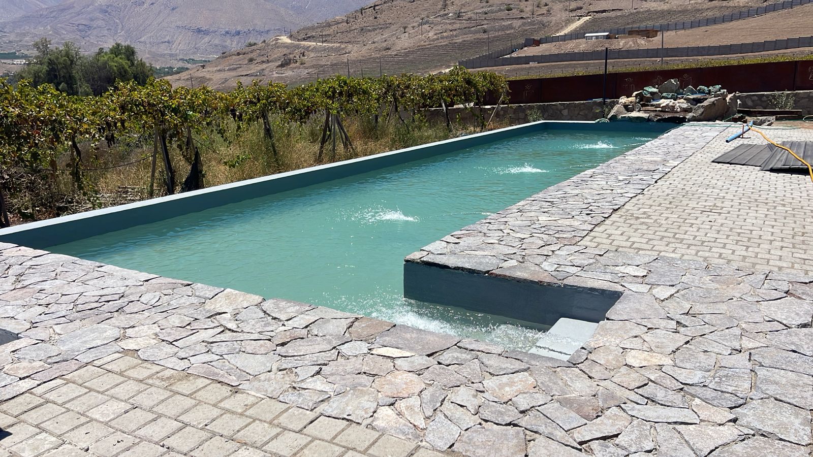 Piscina rectangular con cascada interior, bordes de laja gris y viñedos al fondo